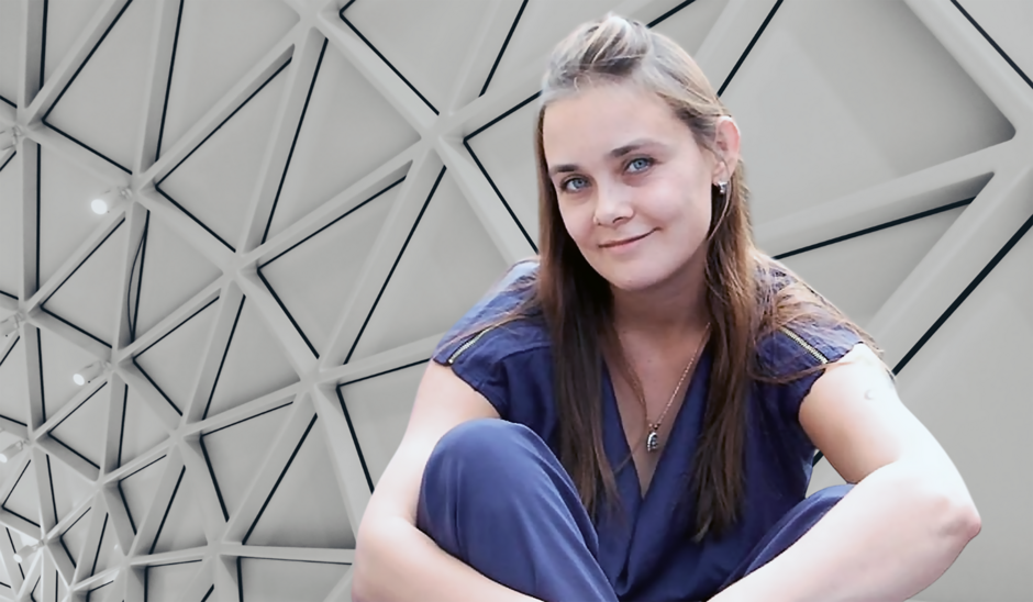 Na zdjęciu kobieta o długich włosach, w koszulce z krótkim rękawem, w tle artystyczna konstrukcja z trójkątów