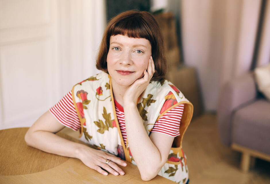 Na zdjęciu kobieta o krótkich włosach, siedząca przy stole, w kwiecistej kamizelce.