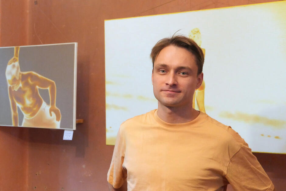 Na zdjęciu mężczyzna w koszulce w odcieniu żółtego, w tle obrazy