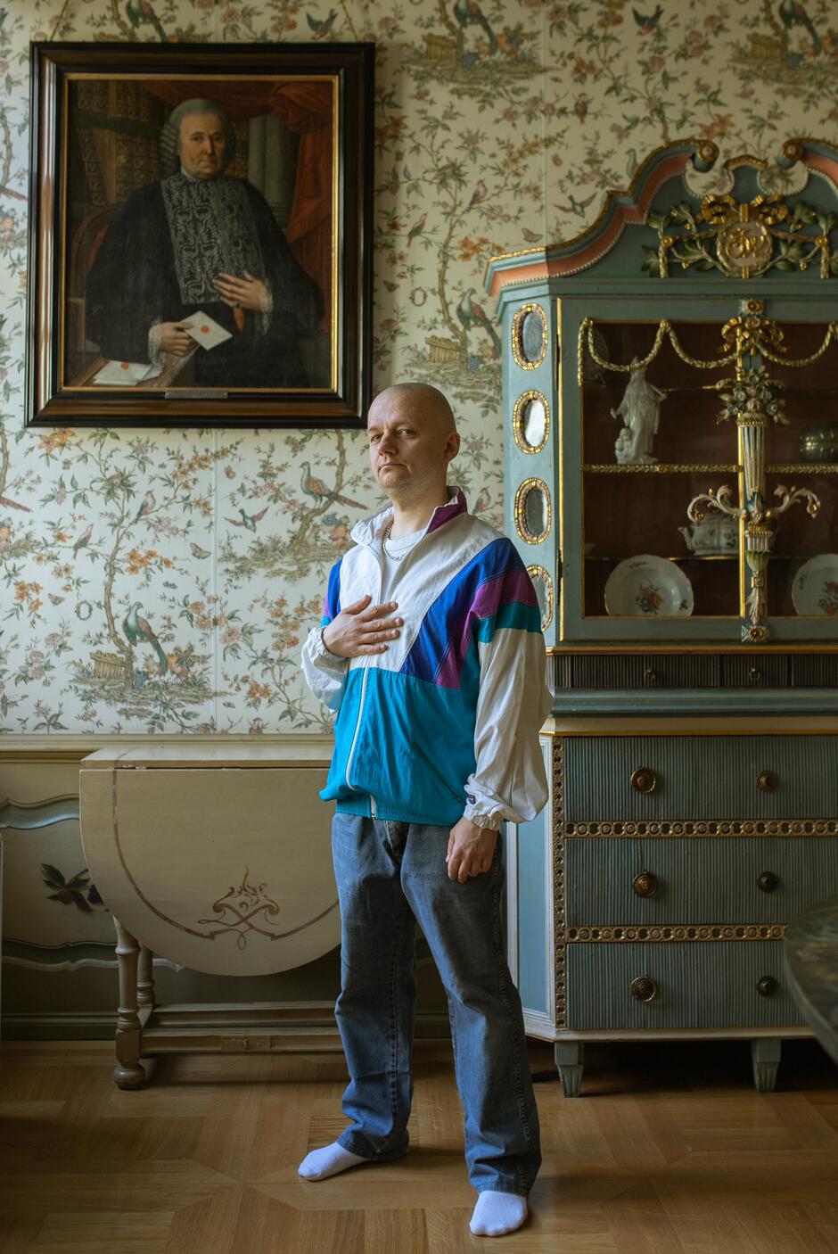 Na zdjęciu mężczyzna w dżinsach oraz niebiesko-białej bluzie w pokoju o wystroju starodawnym, ze zdobioną komodą i obrazem.