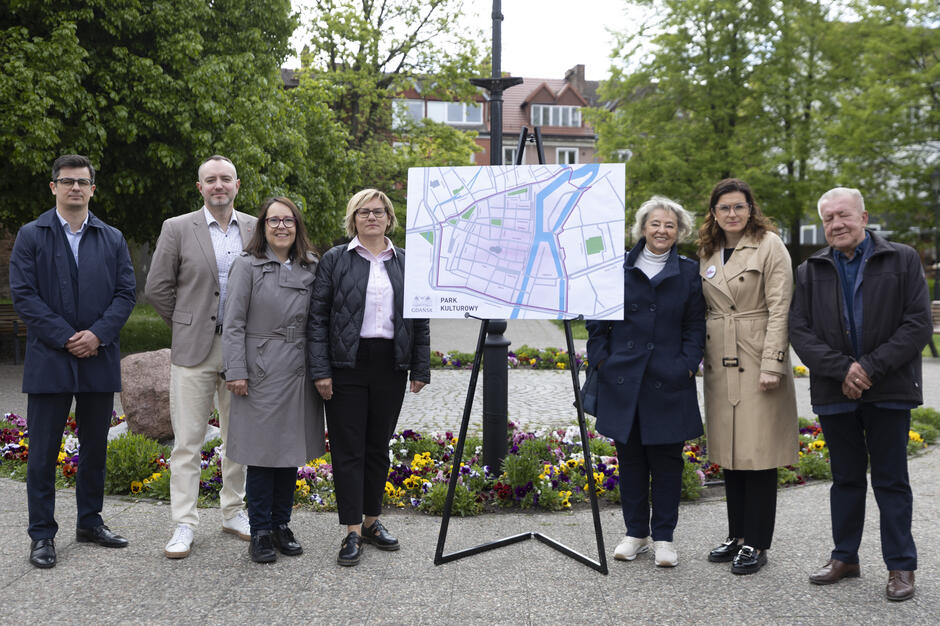 Zdjęcie przedstawia grupę ośmiu osób stojących wokół planszy z mapą, oznaczoną jako Park Kulturowy Gdańsk , co sugeruje prezentację lub spotkanie dotyczące planów urbanistycznych. W tle widoczne są kwiaty, zieleń i budynki mieszkalne, tworząc wiosenną, miejską scenerię.