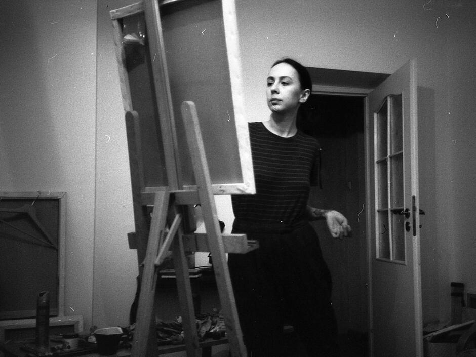 Na zdjęciu kobieta ubrana na czarno, przy sztaludze z płótnem.