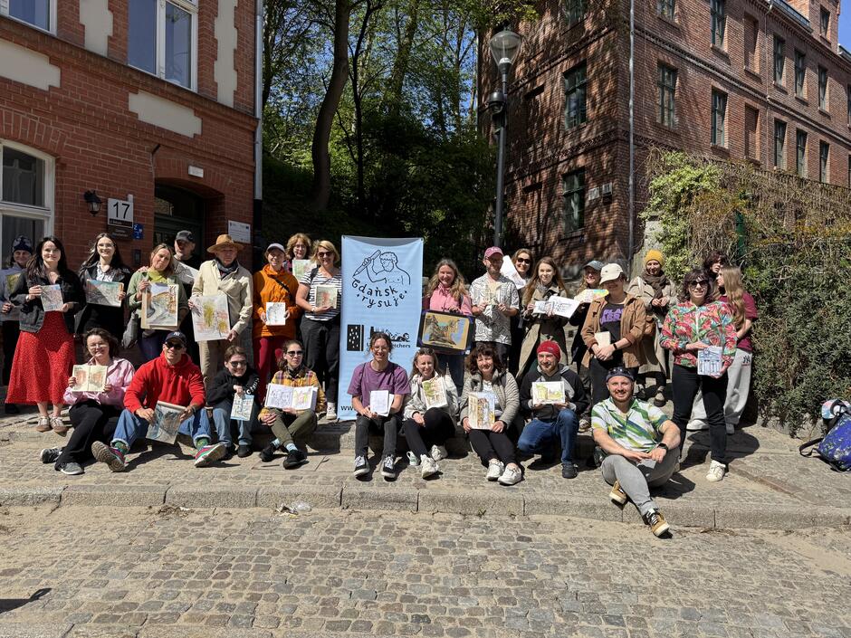 Duża grupa około 30 osób prezentuje swoje rysunki na tle kamienic Biskupiej Górki