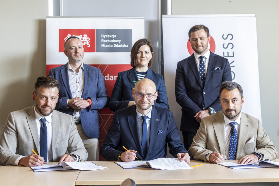 Na zdjęciu widzimy moment podpisania umowy przez trzech mężczyzn w garniturach, siedzących przy stole i trzymających długopisy. W tle stoją trzy osoby – dwóch mężczyzn i jedna kobieta – na tle banerów z logo Dyrekcji Rozbudowy Miasta Gdańska oraz firmy EKOS