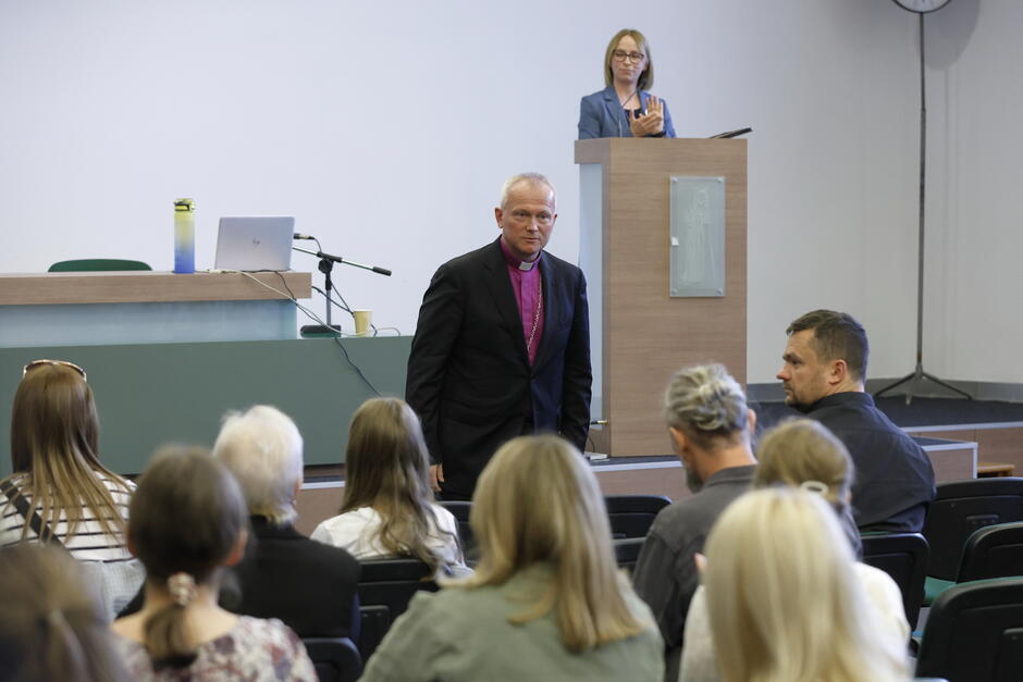 W sali obrad siedzą ludzie, przed nimi stoi luterański biskup, za nim na mównicy stoi kobieta.