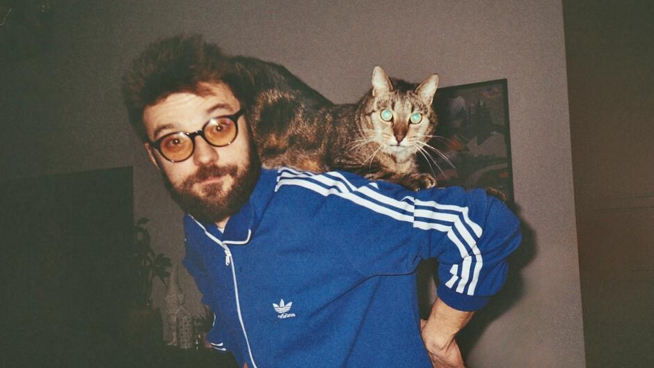 Na zdęciu mężczyzna w niebieskiej bluzie marki adidas, z szarym kotem na lewym ramieniu.