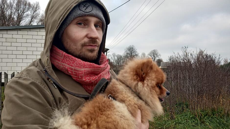 Na zdjęciu mężczyzna w kapturze, w kurtce, trzymający na rękach małego rudawego psa.