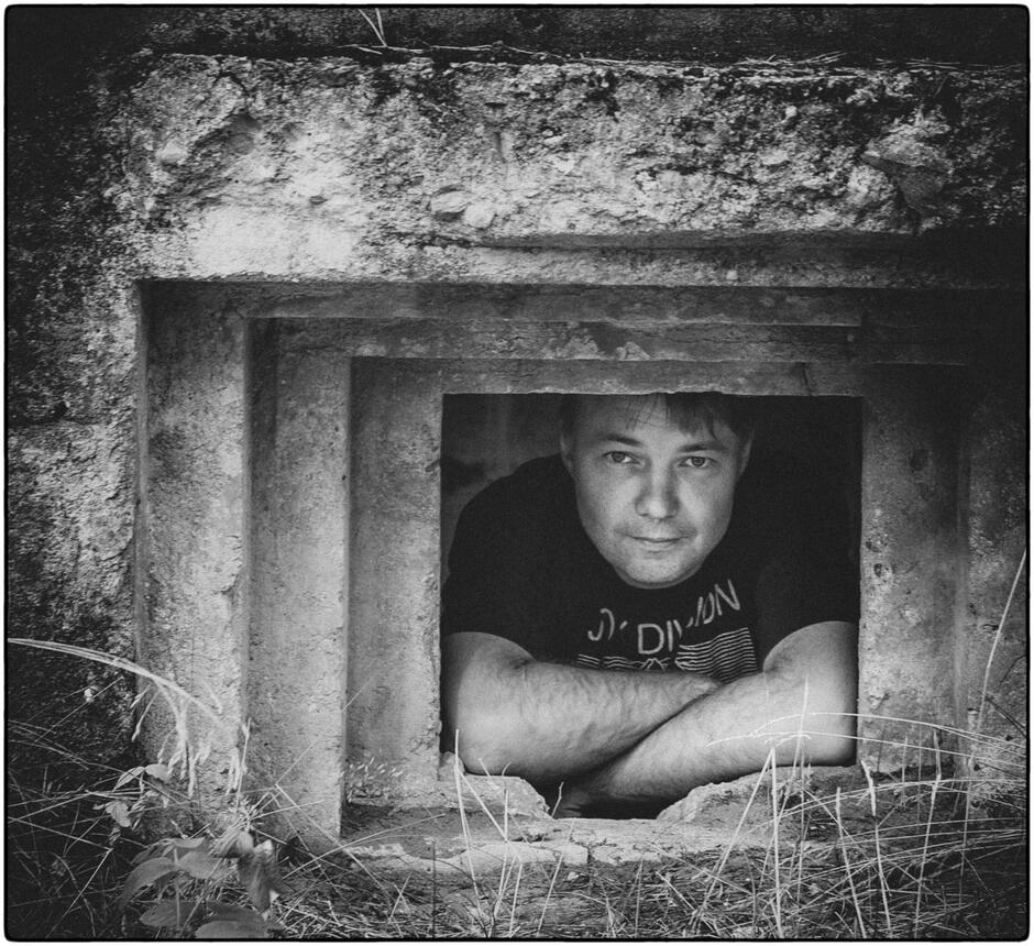 Na zdjęciu mężczyzna z założonymi rękoma, wyglądający z okienka zrujnowanego domu,