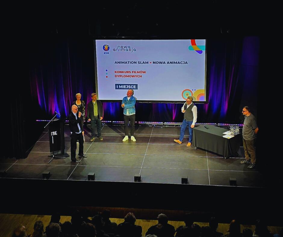 Na zdjęciu widzimy scenę w sali teatralnej lub konferencyjnej, gdzie odbywa się uroczystość wręczenia nagród. Na środku sceny stoi kilku mężczyzn oraz jedna kobieta – prawdopodobnie organizatorzy lub jurorzy wydarzenia. Jeden z mężczyzn przemawia do mikrofonu, co sugeruje, że być może właśnie ogłasza zwycięzcę lub wręcza nagrodę. W tle znajduje się duży ekran z grafiką promującą wydarzenie. Na ekranie widnieje logo „Nowa Animacja” oraz napisy:  „ANIMATION SLAM × NOWA ANIMACJA KONKURS FILMÓW DYPLOMOWYCH I MIEJSCE”