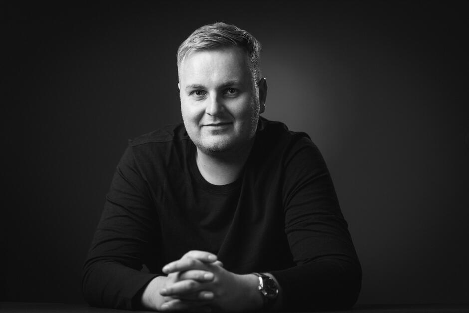 Na zdjęciu mężczyzna o uśmiechniętej twarzy, w czarnej koszulce z długim rękawem.