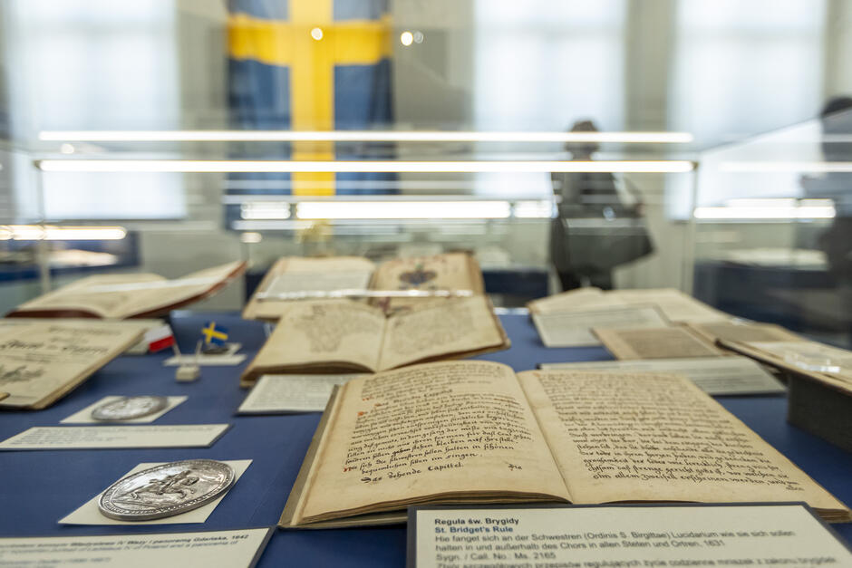 Na zdjęciu widzimy wystawę muzealną lub biblioteczną z cennymi, historycznymi rękopisami i dokumentami. Eksponaty znajdują się w szklanej gablocie, a na pierwszym planie leży otwarta księga zapisana starodawnym pismem. Na etykiecie pod księgą widnieje informacja po niemiecku i angielsku, wskazująca, że jest to Reguła św. Brygidy  – zasady życia zakonnego dla sióstr z Zakonu św. Brygidy, pochodzące z 1631 roku.