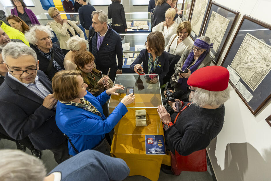 Na zdjęciu widzimy grupę ludzi zgromadzonych wokół gabloty muzealnej podczas wystawy. W środku, za przezroczystą osłoną, znajduje się cenny, prawdopodobnie bardzo stary dokument lub księga. Zwiedzający – w różnym wieku – z dużym zainteresowaniem przyglądają się eksponatowi, wielu z nich robi zdjęcia telefonami lub aparatami.