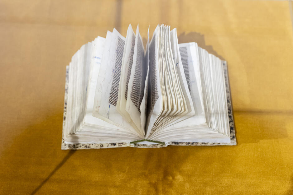 Na zdjęciu widzimy zabytkową, otwartą księgę wystawioną w gablocie na żółtym materiale. Księga ma grube, pergaminowe lub bardzo stare papierowe karty, zapisane gęsto ręcznie pisaną czcionką gotycką lub łacińską. Tekst jest jednolity, ciemny, a marginesy zdobią delikatne, ozdobne inicjały i linie wykonane najprawdopodobniej czerwonym i niebieskim tuszem.