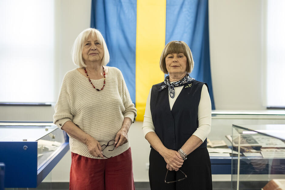 Na zdjęciu widzimy dwie starsze kobiety stojące na tle dużej szwedzkiej flagi (niebieskie tło z żółtym krzyżem), co sugeruje, że znajdują się one na wydarzeniu związanym ze Szwecją — prawdopodobnie wystawie poświęconej relacjom polsko-szwedzkim lub historii Szwecji. Kobiety stoją w otoczeniu gablot muzealnych z dokumentami, manuskryptami i książkami, co dodatkowo potwierdza kontekst wystawienniczy lub muzealny. Obie panie trzymają w dłoniach okulary i patrzą spokojnie w stronę obiektywu. Ich postawa i centralne umiejscowienie na tle flagi sugerują, że mogą być organizatorkami, kuratorkami lub gośćmi honorowymi wydarzenia.