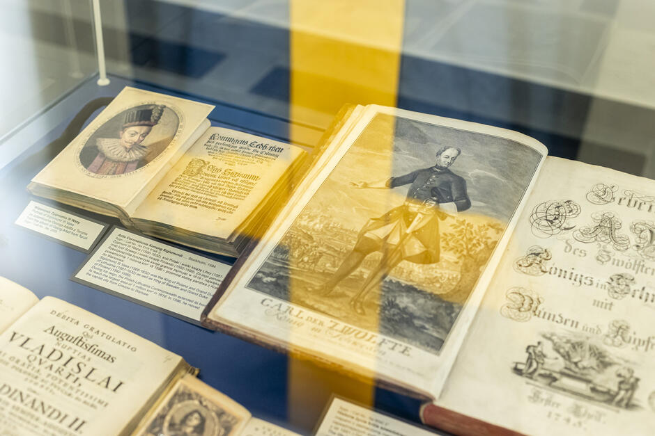 Na zdjęciu przedstawiono ekspozycję zabytkowych książek i druków w muzealnej lub bibliotecznej gablocie. Widoczne są dzieła związane z monarchami Polski i Szwecji – zarówno teksty, jak i portrety. Po lewej stronie widzimy ilustrację portretową króla Zygmunta III Wazy w ozdobnym stroju z charakterystycznym kołnierzem. Obok znajduje się książka zatytułowana „Acta Constitutiones Konunga Sigismundi…” wydana w Sztokholmie w 1587 roku – dokument ten dotyczy panowania Zygmunta III, który był jednocześnie królem Polski i królem Szwecji, co stanowi ważny wątek w historii relacji między tymi dwoma krajami.