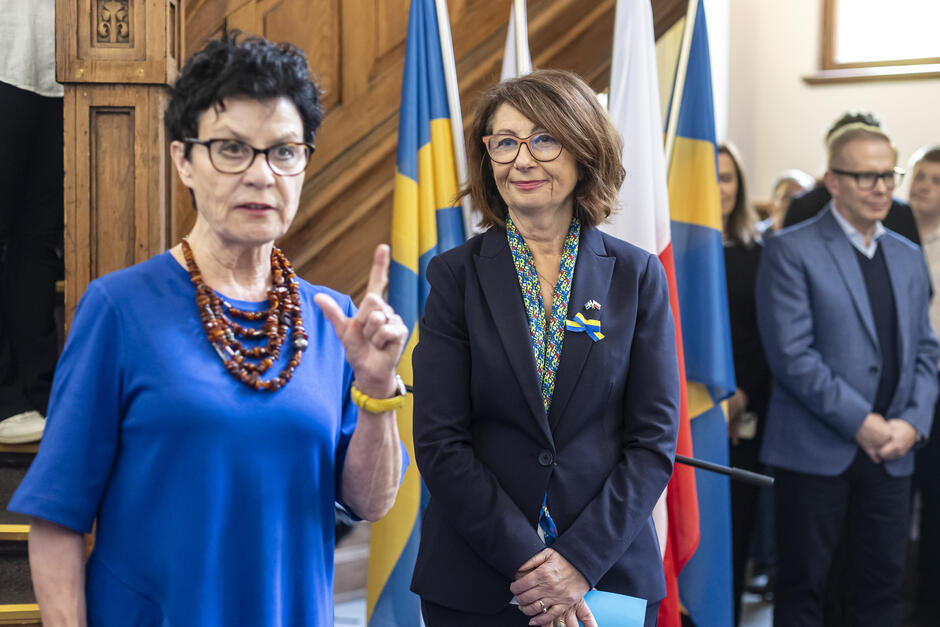 Na zdjęciu widzimy wydarzenie oficjalne, prawdopodobnie otwarcie wystawy lub konferencję związaną z relacjami polsko-szwedzkimi. Na pierwszym planie znajdują się dwie kobiety – jedna z nich przemawia (po lewej stronie, ubrana w niebieską sukienkę i bursztynowy naszyjnik), a druga (po prawej) uśmiecha się, stojąc w tle i mając przypiętą kokardkę w kolorach flag Polski i Szwecji, co wskazuje na temat spotkania.