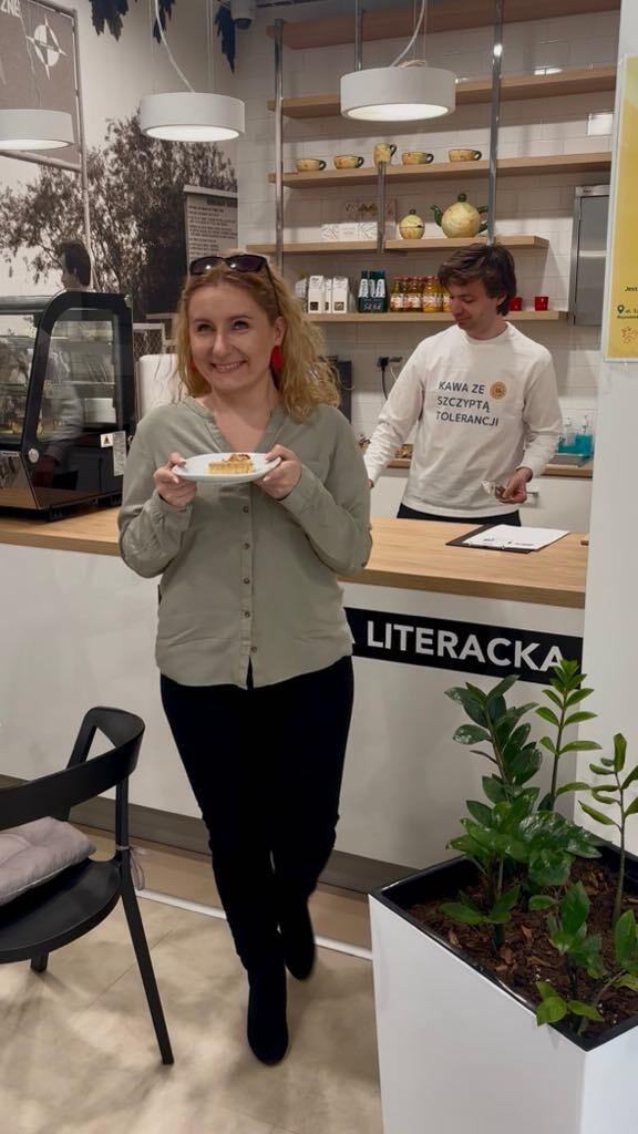 Na zdjęciu uśmiechnięta kobieta trzyma talerzyk z ciastem, stojąc w przytulnej kawiarni o nazwie „Literacka”. W tle barista w koszulce z napisem „Kawa ze szczyptą tolerancji” obsługuje klientów, co tworzy ciepłą i przyjazną atmosferę miejsca.