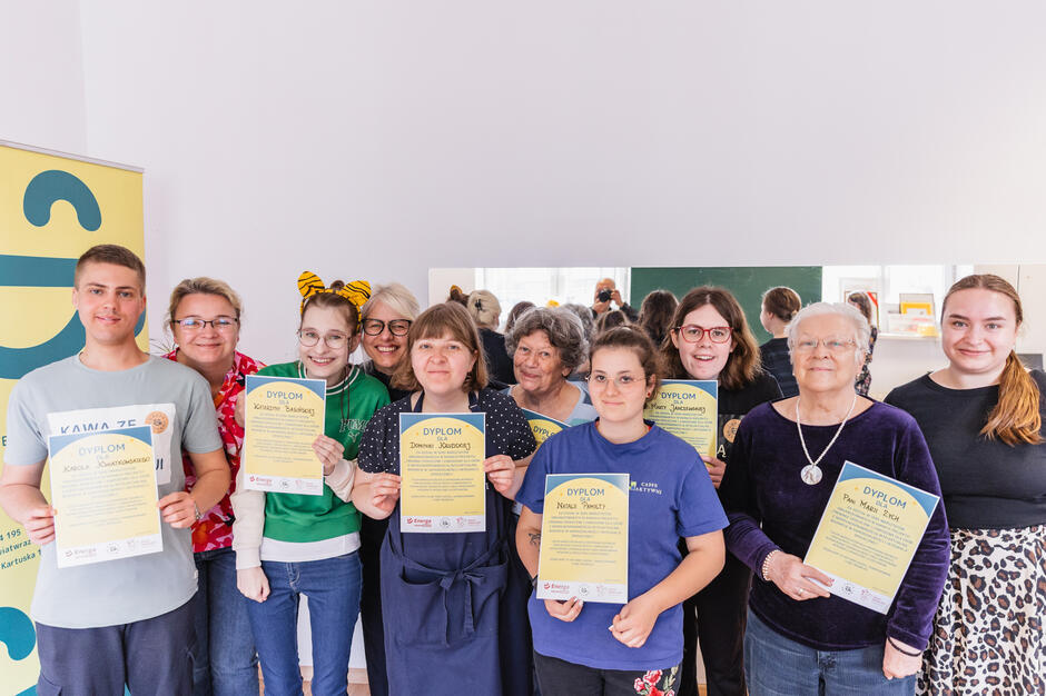 Na zdjęciu widoczna jest grupa uśmiechniętych osób w różnym wieku, trzymających dyplomy — prawdopodobnie uczestników zakończonego projektu lub warsztatów. Atmosfera jest radosna i celebracyjna, co sugeruje udane zakończenie wspólnego przedsięwzięcia.