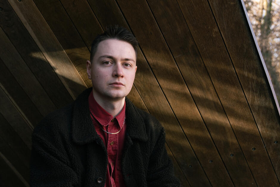 Na zdjęciu mężczyzna w czarnej marynarce patrzący w stronę obiektywu, nad głową konstrukcja z drewna.