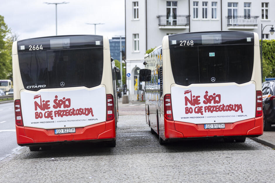 Dwa autobusy stoją zaparkowane na przystanku w mieście, widać je od tyłu. Na autobusach widać hasło namalowane czerwoną czcionką solidarycą : Nie śpij bo cię przegłosują .