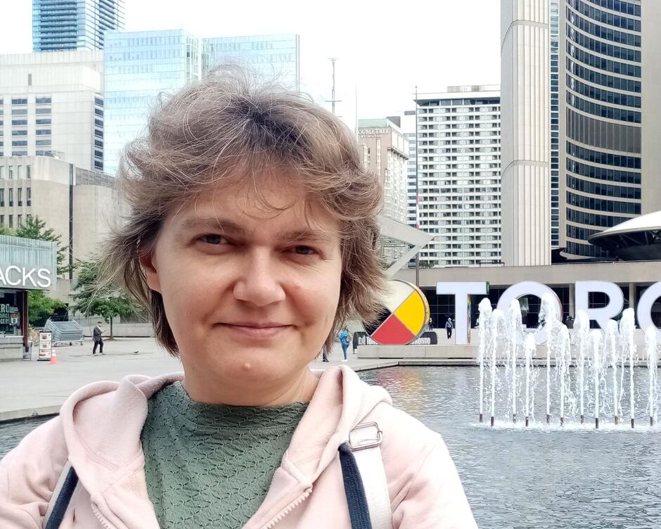 Zdjęcie przedstawia uśmiechniętą twarz kobiety o krótkich włosach, w tle napis i budynki.