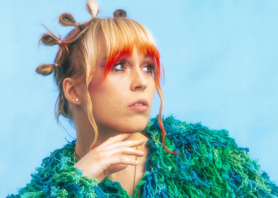 Na zdjęciu kobieta w kolorowej, mechatej bluzie, z fantazyjną fryzurą z blond włosów.