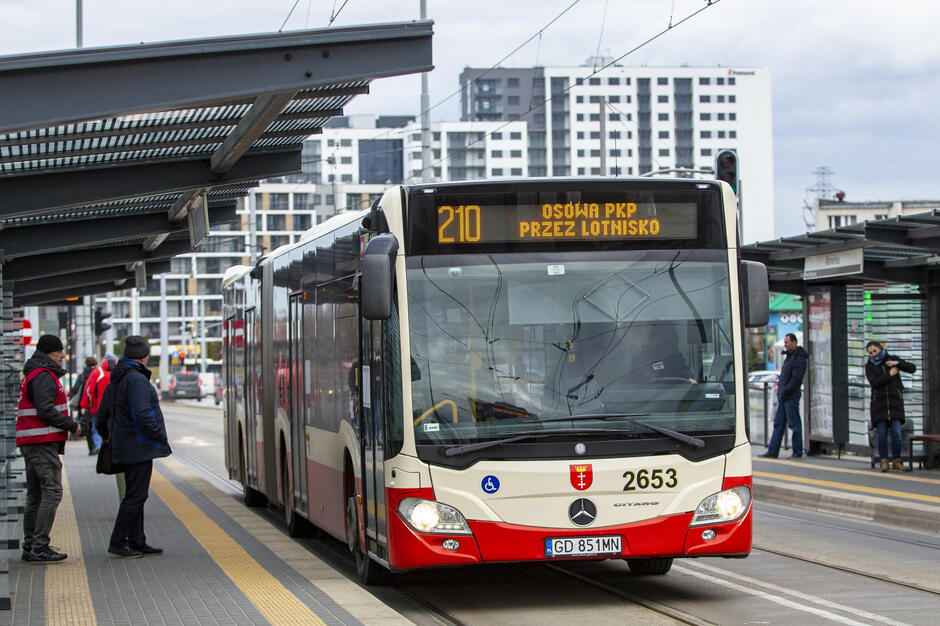 Na zdjęciu widoczny jest autobus linii 210 do Osowej PKP przez lotnisko, stojący na nowoczesnym przystanku w otoczeniu pasażerów i miejskiej zabudowy.