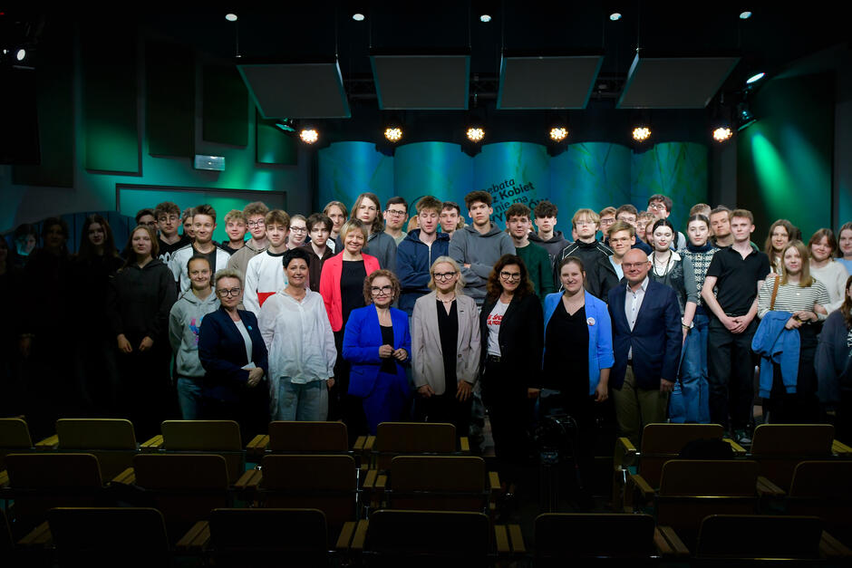 Na zdjęciu widzimy zbiorowe zdjęcie uczestników wydarzenia, które odbyło się w studiu Radia Gdańsk. W tle nadal widoczne jest charakterystyczne turkusowo-niebieskie tło z napisem: „Debata. Bez kobiet nie ma wyborów” oraz symbolem kobiety.