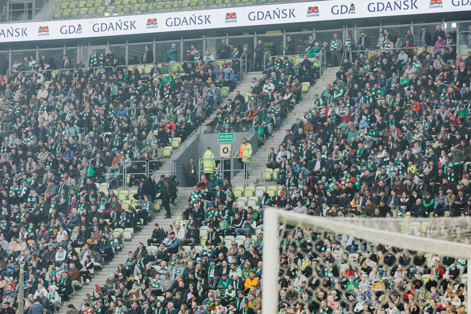 duży tłum ludzi na stadionie.Tłum składa się zarówno z mężczyzn, jak i kobiet, wszyscy w zielono-białych paskowych koszulach.Stoją w rzędach zielonych foteli i skierowani są w kierunku prawej strony jest bramka z siatkę przed nią.Ponad tłumem wiszące z sufitu banery z napisem GDANSK i GDAńSK stoi również kilka osób stojących na schodach prowadzących do stoisk.Wydaje się, że to mecz piłki nożnej lub mecz piłkarski.