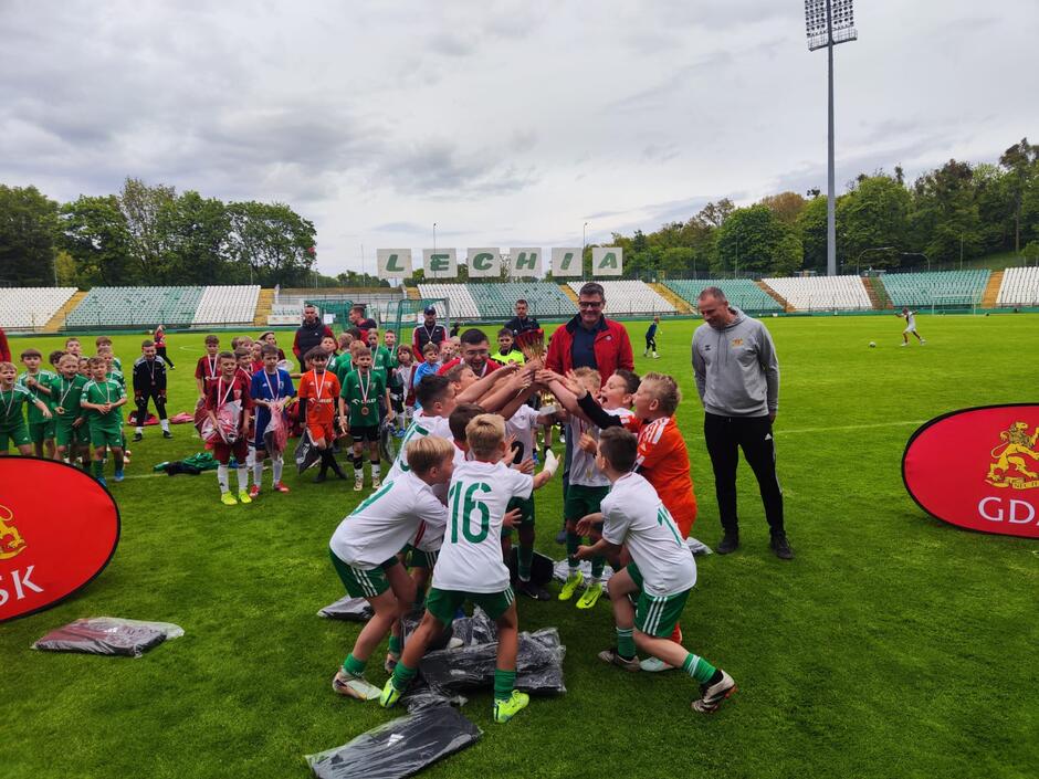 Drużyna młodych piłkarzy świętuje zwycięstwo, wznosząc puchar na stadionie Lechii Gdańsk podczas ceremonii wręczenia medali.