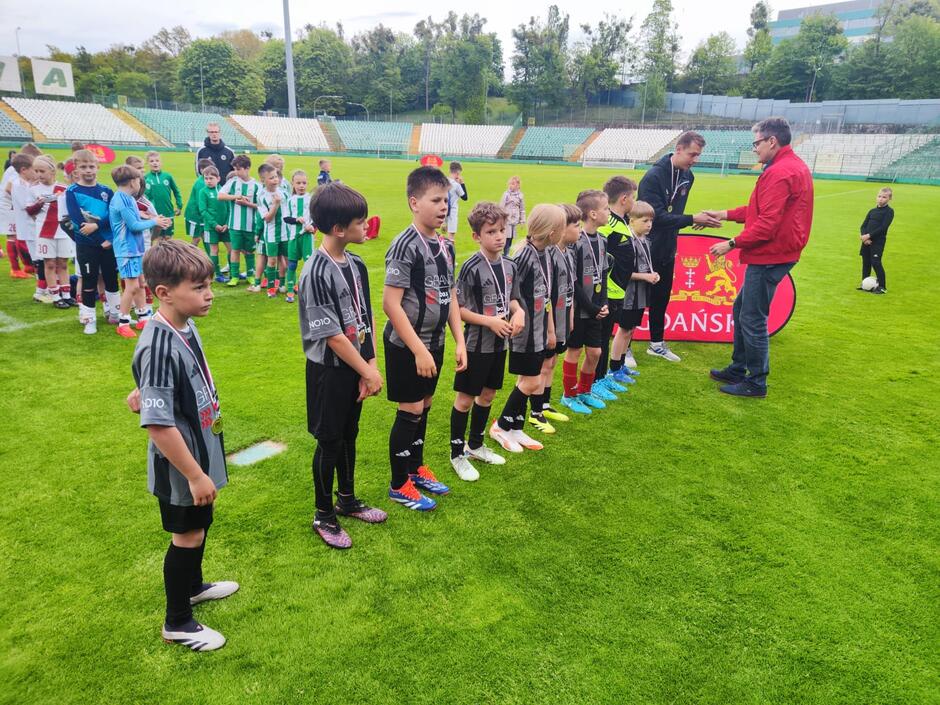 Młodzi piłkarze w czarnych strojach odbierają medale podczas ceremonii wręczenia nagród na stadionie, a w tle stoją inne drużyny.