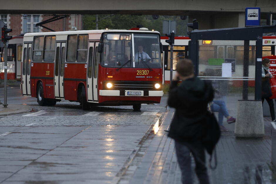 Klasyczny czerwono-biały autobus Ikarus wjeżdża na przystanek w deszczowy dzień, mijając ludzi oczekujących pod wiatą. To nostalgiczny widok nawiązujący do dawnego taboru komunikacji miejskiej