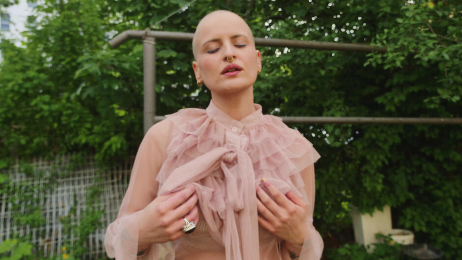 Na zdjęciu kobieta bez włosów, w sukni z delikatnego materiału, na palcu duży czarny pierścień, w tle zieleń i trzepak.