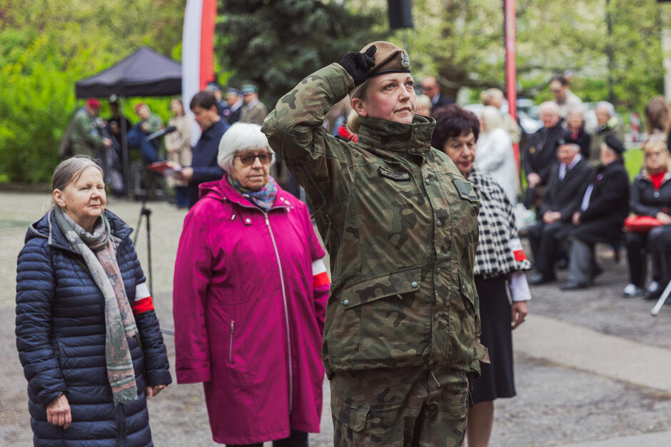 Zdjęcie ukazuje uroczystość patriotyczną, podczas której kobieta w mundurze wojskowym oddaje honory, stojąc na czele grupy uczestników – w tym seniorek z opaskami biało-czerwonymi. W tle widoczna jest zgromadzona publiczność, co nadaje scenie podniosły i uroczysty charakter.

