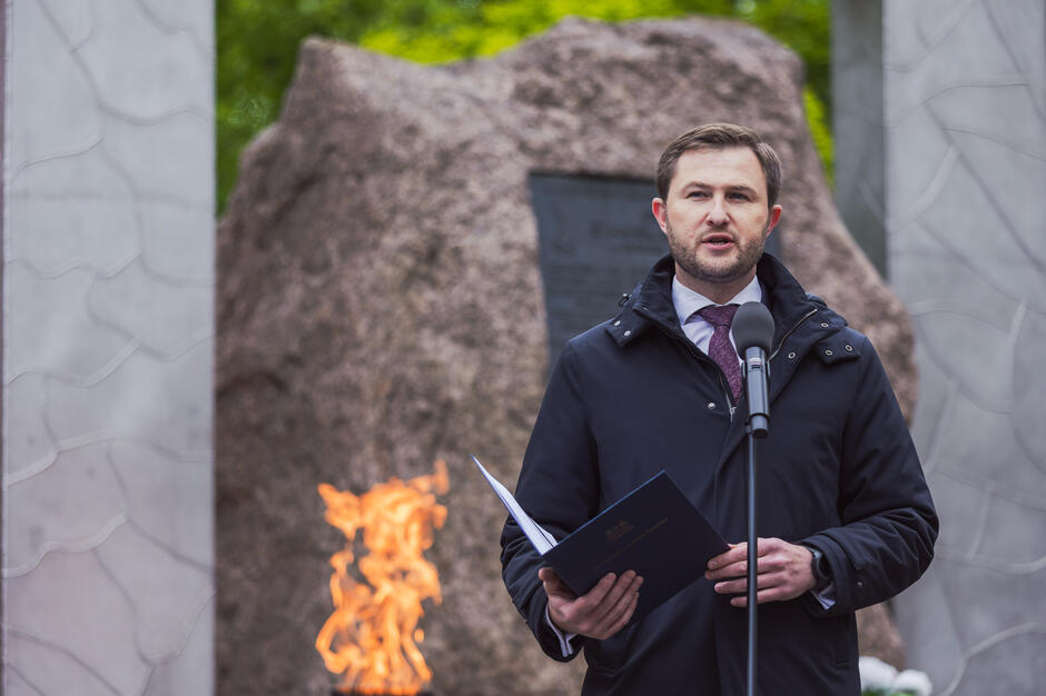 Zdjęcie przedstawia mężczyznę w garniturze i kurtce, przemawiającego przy mikrofonie podczas oficjalnej uroczystości patriotycznej. W tle widoczny jest pomnik z płonącym zniczem oraz tablicą pamiątkową, co nadaje wydarzeniu podniosły i uroczysty charakter. 