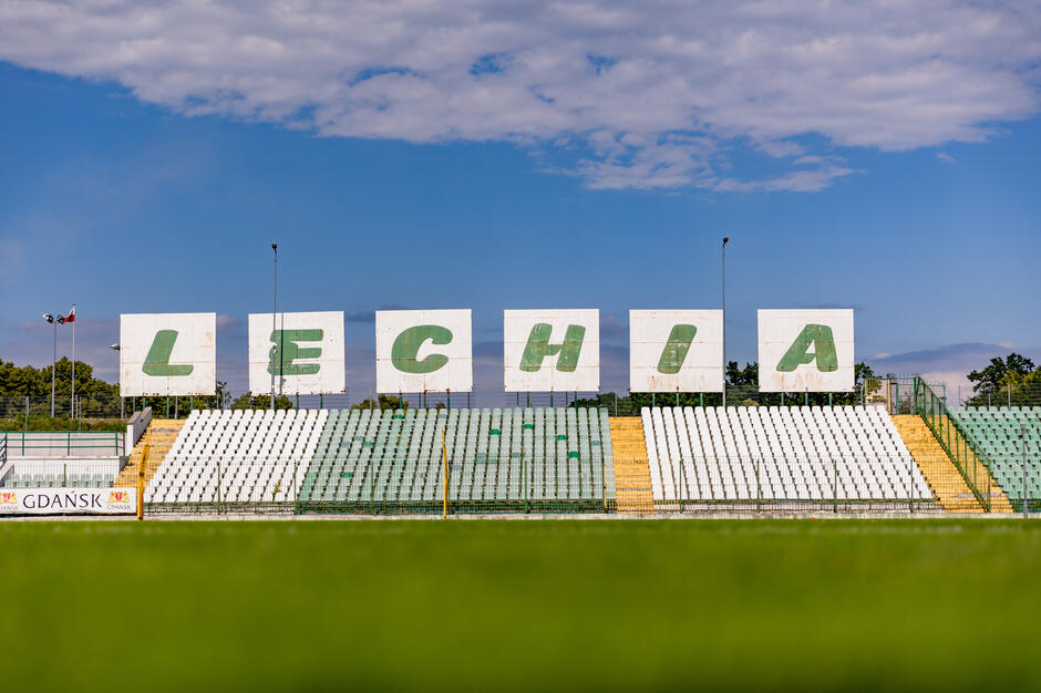 duży znak, który czyta LEchia w białych literach na niebieskim tle nieba. znak znajduje się na pustym stadionie z rzędami białych foteli skierowanych w kierunku prawej strony jest napisany odważną, bezszeryfową czcionką.