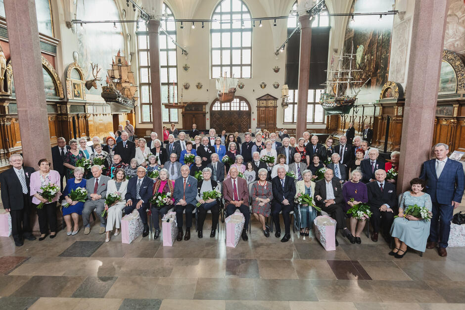 Uroczyste, grupowe zdjęcie seniorów, którzy wzięli udział w ceremonii jubileuszu długoletniego pożycia małżeńskiego. W tle widać zabytkowe wnętrze sali z makietami statków i historycznymi malowidłami.