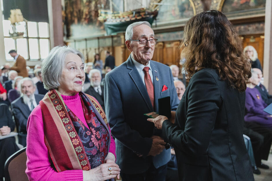 Starsze małżeństwo odbiera medal od przedstawicielki władz miasta podczas oficjalnej ceremonii. Na twarzach widać radość i dumę z otrzymanego wyróżnienia.