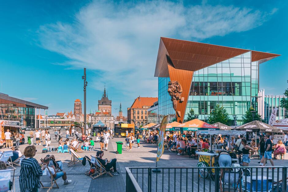 To zdjęcie przedstawia tętniącą życiem przestrzeń miejską przy Forum Gdańsk, z widokiem na historyczną panoramę Starego Miasta Gdańska. W centrum kadru widoczna jest nowoczesna architektura z charakterystycznym budynkiem z przeszkloną fasadą i rzeźbą lwa – to część Forum Gdańsk.