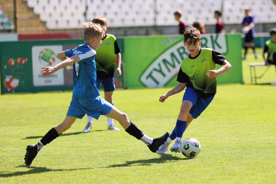 Dwóch młodych chłopców rywalizuje o piłkę podczas meczu piłkarskiego na boisku trawiastym.