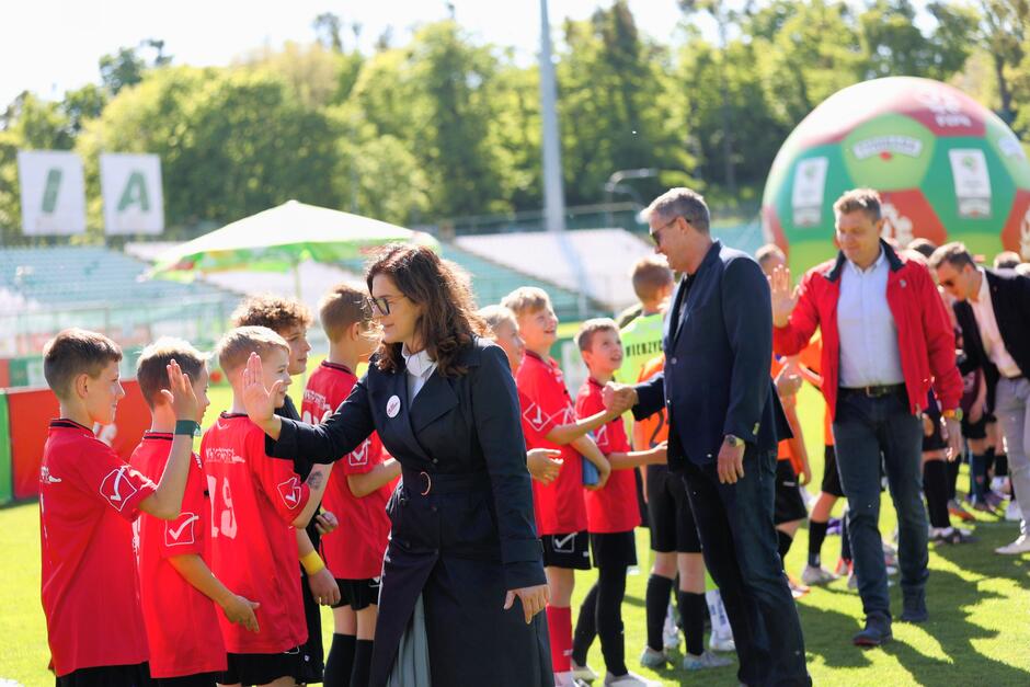 Dorośli witają się i przybijają piątki z młodymi piłkarzami ustawionymi w szeregu na boisku.