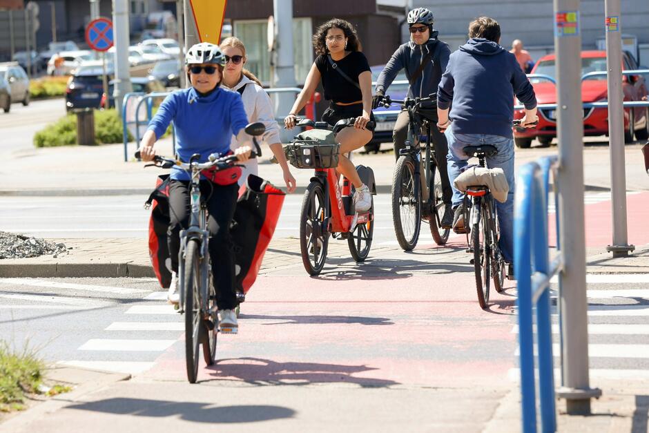Na zdjęciu widać grupę osób jadących na rowerach po wyznaczonej ścieżce rowerowej w miejskim otoczeniu. Pogoda jest słoneczna, a rowerzyści są ubrani lekko, co sugeruje, że to ciepły dzień.