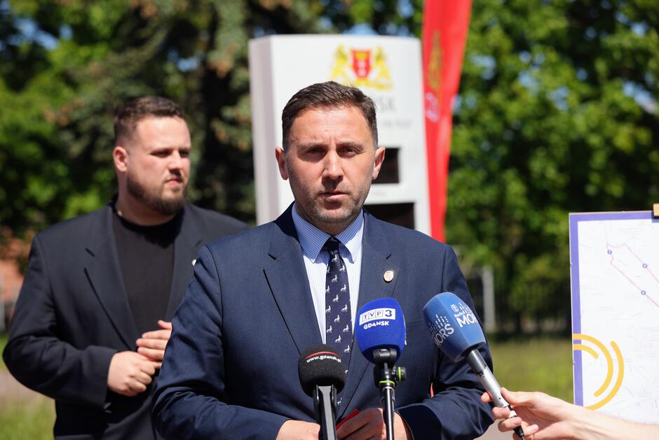 Na zdjęciu widać mężczyznę w garniturze, który przemawia przed mikrofonami podczas konferencji prasowej na świeżym powietrzu. W tle stoi inny mężczyzna oraz widoczna jest tablica z herbem i mapa, co sugeruje oficjalne wydarzenie związane z samorządem lub infrastrukturą miejską.