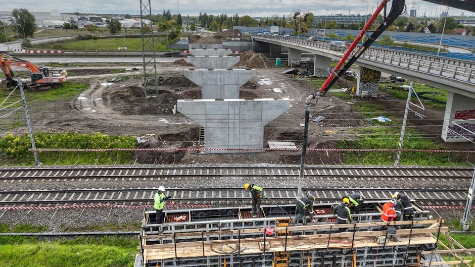 na zdjęciu plac budowy, na dole zdjęcia widać pracujących robotników, za nimi w tle fragment nowych podpór betonowych
