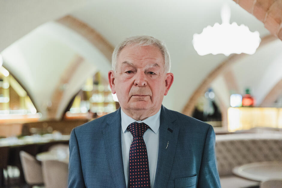 Starszy mężczyzna stoi wyprężony i patrzy prosto w obiektyw pozując do fotografii. Stoi w sali restauracyjnej w starym budynku z łukowym sklepieniem.
