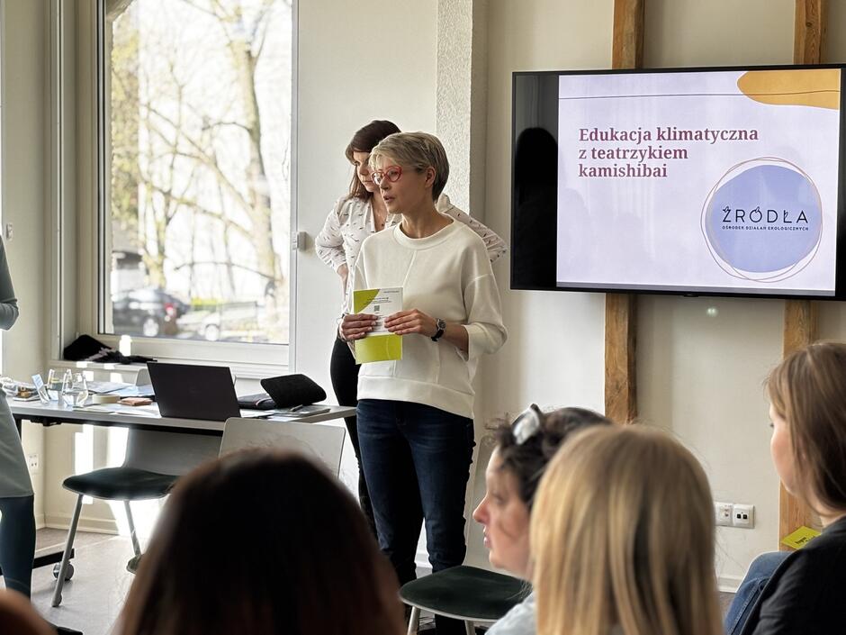 Na zdjęciu widzimy prowadzącą prezentację kobietę podczas szkolenia dla nauczycieli, którego tematem jest „Edukacja klimatyczna z teatrzykiem kamishibai”, jak wskazuje slajd wyświetlany na ekranie. Uczestnicy siedzą i słuchają z zainteresowaniem, a całość odbywa się w jasnym, nowoczesnym pomieszczeniu.