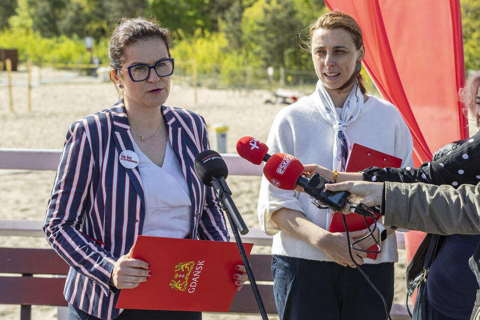 grupa ludzi zebrała się wokół kobiety, która trzyma czerwony folder z logo na nim, kobieta ma na sobie pasiastą bluzę i okulary i wydaje się rozmawiać z mediami, przed nią kilka mikrofonów, wskazujących, że jest przesłuchana, mężczyzna w garniturze i krawatie stoi po jej prawej stronie, trzymając folder i przemawiając do jednego jest na wydarzeniu publicznym lub konferencji prasowej.