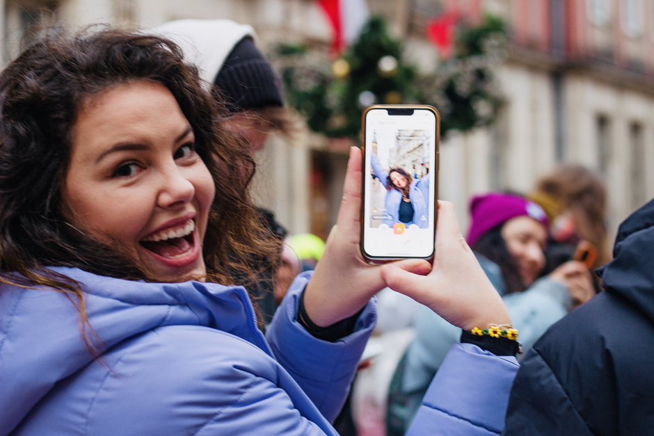 Młoda kobieta w niebieskiej kurtce z radością pozuje do zdjęcia, trzymając telefon, na którym widać jej ujęcie z tej samej chwili. 