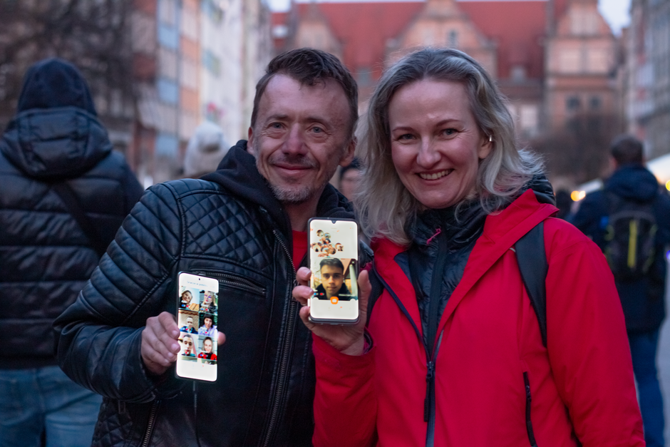 Para uśmiechniętych osób pozuje na ulicy, trzymając w dłoniach smartfony z widocznymi zdjęciami i wideorozmowami. 