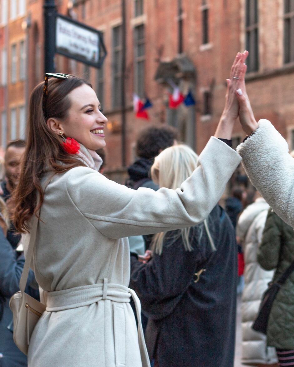 Dwie uśmiechnięte kobiety w płaszczach dają sobie piątkę  na tętniącej życiem ulicy.
